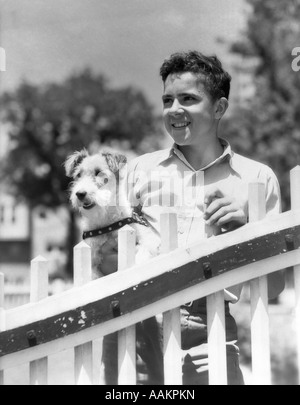 1930ER JAHREN TEENBOY LÄCHELND DURCH EINEN ZAUN HOLDING WIRE FOX TERRIER HUND STEHEND Stockfoto