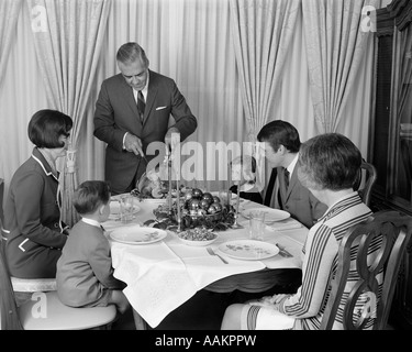 1960ER JAHREN GROßVATER STEHEN CARVING THANKSGIVING-TRUTHAHN, UMGEBEN VON DREI GENERATIONEN-FAMILIE Stockfoto