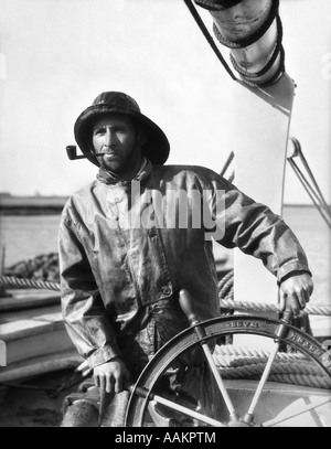 1920S 1930S MANN ALTE SALZ SAILOR FISCHER AN DER SPITZE DER FISCHERBOOT TRAGEN VON SCHLECHTEM WETTER GEAR UND RAUCHT EINE PFEIFE Stockfoto