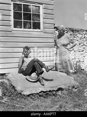 1940ER JAHRE BAUERNJUNGE GELEHNT GEBÄUDE PFEIFE MIT GROßMUTTER SAH UM ECKE IHN MIT HAND ON HIP Stockfoto