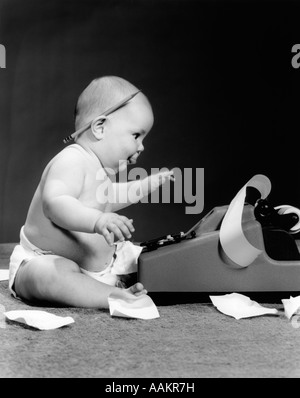 1960ER JAHREN SEITENANSICHT DES MOLLIG BABY SITZT HINTER RECHENMASCHINE MIT BLEISTIFT HINTER DEM OHR & BAND AUF BODEN VERSTREUT Stockfoto