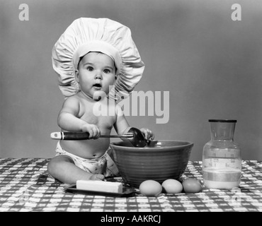 1960ER JAHRE BABY SITZEND AUF KARIERTEN TISCHDECKE TRAGEN KOCHMÜTZE MISCHEN EIERN MILCH & MEHL IN EINER GROßEN SCHÜSSEL BLICK IN DIE KAMERA Stockfoto