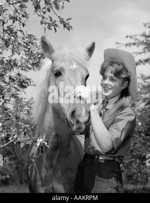 1960ER JAHRE TEEN GIRL IN COWBOY-HUT-PETTING-PONY UNTER DEN BÄUMEN Stockfoto