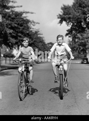 1930S 1940S ZWEI JUNGEN REITEN FAHRRÄDER AM BAUM GESÄUMTEN STRAßE Stockfoto