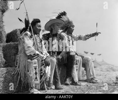 1940ER JAHREN PAAR DER INDIANER IN VOLLER TRACHT AUF STROHBALLEN SITZEND MIT COWBOY ZWISCHEN IHNEN Stockfoto