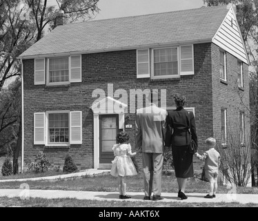 1950ER JAHRE RÜCKSEITE DES STEHEN VOR NEUEN DOMIZIL HAND IN HAND 4-KÖPFIGE FAMILIE Stockfoto