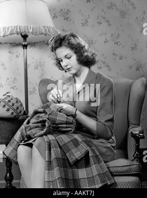 1940ER JAHRE JUNGE FRAU TEENAGER SITZEN IM STUHL UNTER LAMPE NÄHEN Stockfoto