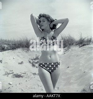 1950ER JAHRE BRÜNETTE BADENIXE IN POLKA DOT BIKINI IN SAND MIT HÄNDE HINTER KOPF STEHEND Stockfoto