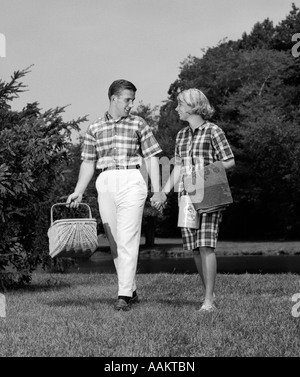 1960ER JAHRE TEENAGER-PAAR HAND IN HAND GEHEN MIT PICKNICK-KORB, REDEN, FLIRTEN, DATING Stockfoto