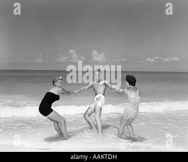 1950ER JAHRE MANN UND ZWEI FRAUEN TUMMELN IN BEACH SURF Stockfoto