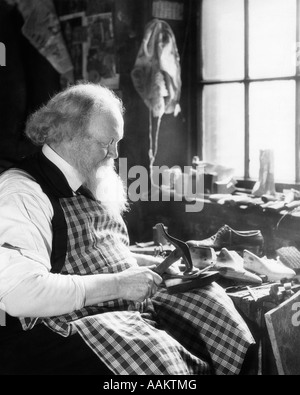 1920S 1930S BÄRTIGEN ÄLTEREN SCHUSTER DURCH FENSTER AN WERKBANK REPARATUR EINEN SCHUH TRÄGT EINE AUFGEGEBENE SCHÜRZE UND HÄLT EINEN HAMMER Stockfoto