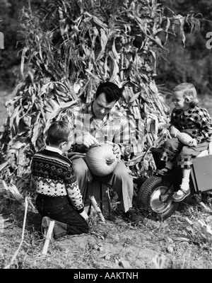 1950ER JAHRE JUNGE & MÄDCHEN SITZEN VOR MAISSTROH BEOBACHTEN VATER KÜRBIS SCHNITZEN Stockfoto