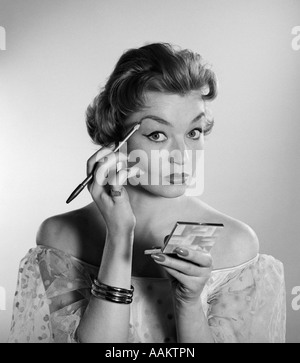 1950S 1960S WOMAN LOOKING AT KAMERA ANWENDUNG MAKE-UP EYE BROW BLEISTIFT HOLDING TASCHENSPIEGEL Stockfoto