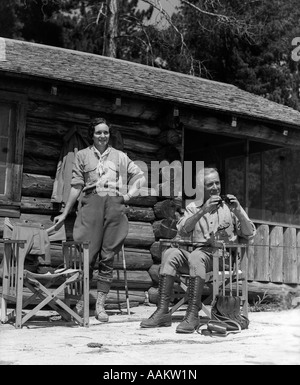 1930ER JAHREN PAAR MANN FRAU VERANDA BLOCKHAUS FRAU STEHT VOM STUHL ÄLTERER MANN SITZT HALT FERNGLAS URLAUB Stockfoto