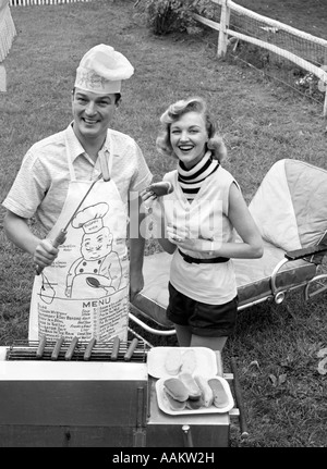 1950ER JAHREN PAAR HINTERHOF GRILL KOCHEN HOTDOGS MANN TRÄGT SCHÜRZE TOQUE & AUFGESPIEßT HOT-DOG Stockfoto