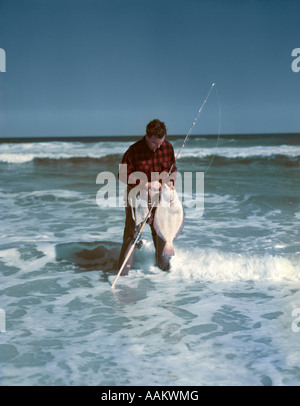 1950ER JAHRE MANN ROT SCHWARZ ÜBERPRÜFT SHIRT STAND IN DER BRANDUNG ENTFERNEN FLUNDER VOM HAKEN HAKEN SURF CASTING SPORT FISCHEN OZEAN Stockfoto