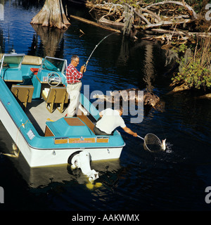 1970ER JAHREN ZWEIER ÄLTERER MÄNNER ANGELN IN CYPRESS SWAMP AUS BLAU WEIßE MOTORBOOT AUßENBORDER EINEN MANN ROD REEL ANDERE NETZE NET FISCH Stockfoto