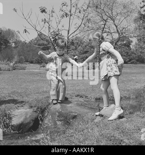 1950ER JAHRE MÄDCHEN HELFEN JÜNGEREN KNABEN ÜBER BACH Stockfoto