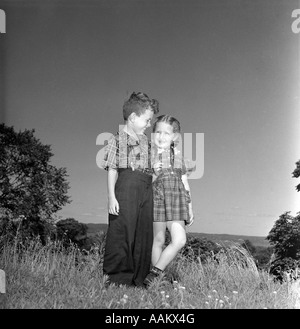 1940ER JAHREN LÄCHELND BOY KARIERTEN HEMD UND MÄDCHEN KARIERTEN KLEID ZUSAMMEN NEBENEINANDER AUF HÜGEL WIESE STEHEN Stockfoto