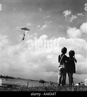 1940ER JAHRE JUNGES MÄDCHEN UND JUNGEN STEHEN NEBENEINANDER FLIEGEN PATRIOTISCHE FLAGGE KITE ÜBER RIVER Stockfoto