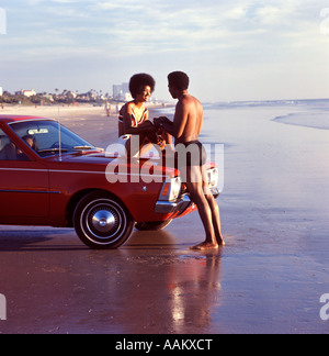 1970ER JAHREN LÄCHELND AFROAMERIKANISCHE ROMANTIKURLAUB ZU ZWEIT AM STRAND FRAU SITZT AUF DEM AUTO KAPUZE MANN HAND IN HAND Stockfoto