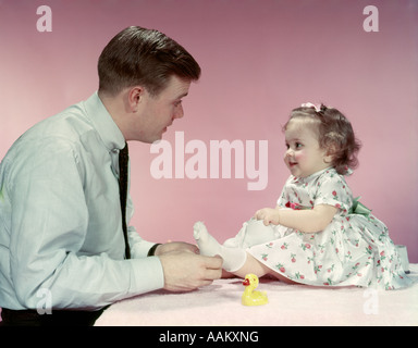 1950ER JAHREN VATER MANN KLEINES MÄDCHEN IN PRINT GEBLÜMTEN KLEID ROSA UND WEIßEN ELTERN TOCHTER DRESSING SOCKEN ANZIEHEN Stockfoto