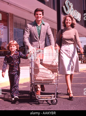 1960ER JAHREN 3-KÖPFIGE FAMILIE VATER SCHIEBEN LEBENSMITTELGESCHÄFT WARENKORB AUF PARKPLATZ Stockfoto