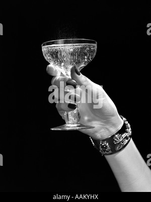 1930ER JAHREN 1940ER JAHRE 1950ER JAHRE FRAU HAND VERZIERTEN METALLARMBAND HOCHHALTEN SILVESTER TOAST GLAS CHAMPAGNER VOR SCHWARZEM HINTERGRUND Stockfoto