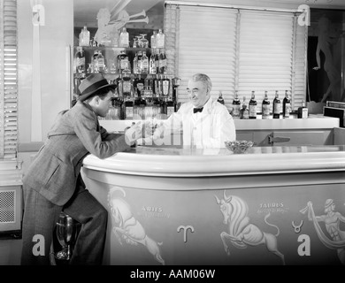 1930ER JAHREN 1940ER JAHRE 1950ER JAHRE MANN STÜTZTE SICH AUF BAR BARKEEPER EINEM ANDEREN ALKOHOLISCHES GETRÄNK BESTELLEN, IM ART-DECO-STIL COCKTAIL LOUNGE Stockfoto