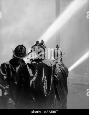 1920ER JAHRE RÜCKANSICHT DER VIER FEUERWEHRLEUTE IN SCHWARZ SLICKERS SPRITZEN ZWEI SCHLÄUCHE Stockfoto