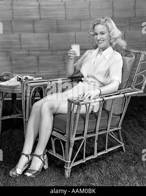 1940ER JAHRE BLONDE FRAU IN BAMBUS STUHL HÄLT GLAS MILCH TRAGEN WEIßE HOSE, BLICK IN DIE KAMERA LÄCHELN Stockfoto