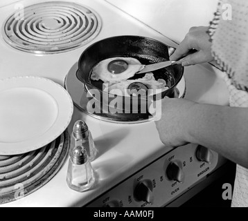 1940S 1950S HÄNDE FRAU BRATEN EIERN IN EINER EISERNEN PFANNE BEI ELEKTRISCHEN HERD SALZ- UND PFEFFERSTREUER Stockfoto