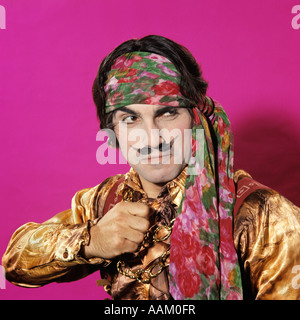 CHARAKTER MANN LUSTIGE AUSDRUCK GESTE GEKLEIDET IN PIRATE KOSTÜM GEFÄLSCHTE SCHNURRBART ROSA HINTERGRUND GOLD SEIDENHEMD STIRNBAND Stockfoto