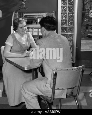 1950ER JAHRE TEEN PAAR SITZT AM TISCH IM DINER JUKEBOX IM HINTERGRUND LÄCHELT INDOOR Stockfoto