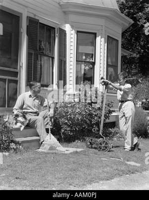 1940ER JAHRE HARKEN VATER SITZT AUF VERANDA SCHRITTE STREICHELN HUND HALTEN IM GESPRÄCH MIT SOHN STAND HALTEN RASENMÄHER Stockfoto