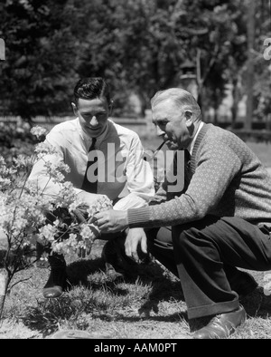 1930ER JAHREN 1940ER JAHREN 2 ERWACHSENE MÄNNER VATER & SOHN KNIEND IM GARTEN BLICK AUF BLÜHENDER STRAUCH Stockfoto