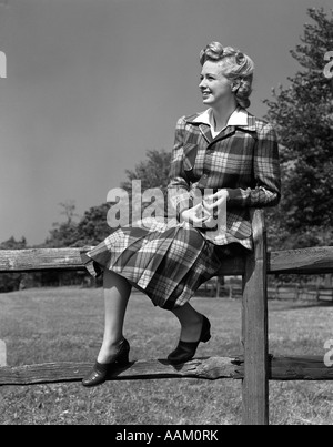 1940ER JAHRE FULL LENGTH PORTRAIT OF BLONDE FRAU SITZT AUF EINEM HÖLZERNEN ZAUN TRAGEN KARIERTE TARTAN ANZUG Stockfoto