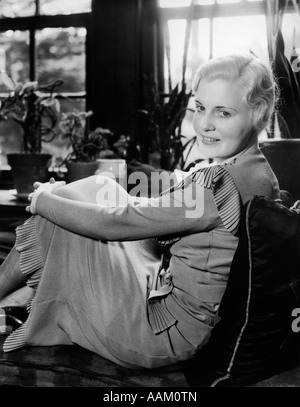 1920S 1930S BLONDE FRAU LÄCHELND MIT SITZEN AUF DEN KNIEN IN FENSTERPLATZ MIT BLICK IN DIE KAMERA ARME Stockfoto