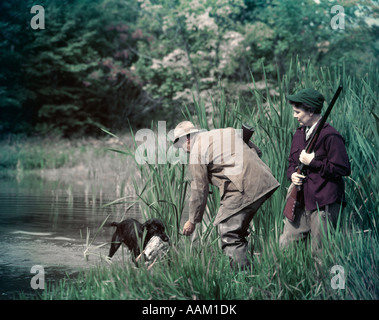 1950ER JAHRE MANN FRAU PAAR SCHROTFLINTE SCHIEßEND JAGD DURCH SEE TEICH SCHWARZER HUND LABRADOR RETRIEVER WIEDER WASSER GEFLÜGEL ENTE Stockfoto