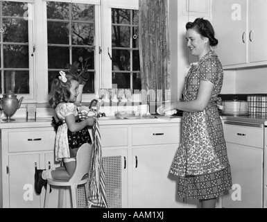 1930S 1940S FRAU MUTTER MIT TOCHTER DER MÄDCHEN KNIEND AUF STUHL HILFT MIT WASCHEN UND TROCKNEN GESCHIRR IN DER KÜCHE Stockfoto