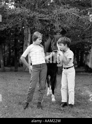 1970ER JAHRE JUNGE & MÄDCHEN DRAUßEN HOLDING PONY VON ZAUMZEUG Stockfoto