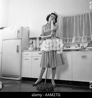 1950ER JAHREN GELANGWEILT FRAU HAUSFRAU TRÄGT SCHÜRZE STÜTZTE SICH AUF MOP AUF KÜCHENBODEN Stockfoto