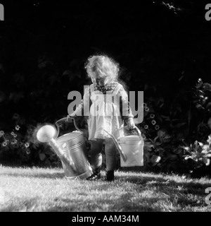 1930ER JAHRE NIEDLICHE KLEINE BLONDE MÄDCHEN IN PINAFORE HOLDING ZWEI TRÄNKEN DOSEN WANDERN IM HINTERHOF Stockfoto