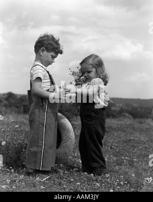 1940S 1950S KLEINES MÄDCHEN ÜBERGABE GÄNSEBLÜMCHEN ZU JUNGE STAND IM BEREICH DER BLUMEN Stockfoto