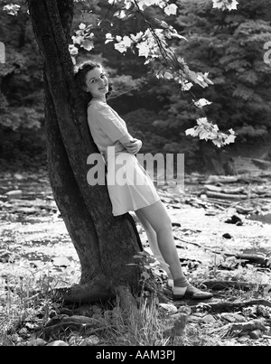 1940ER JAHREN LÄCHELND JUNGE FRAU TRÄGT KURZEN ROCK UND SCHUHE SATTEL GELEHNT BAUM PER STREAM Stockfoto