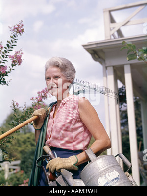 1960ER JAHRE SENIOR FRAU GÄRTNER HALTEN RAKE SCHLAUCH GIEßKANNE SCHAUFEL WERKZEUGE MIT HANDSCHUHEN Stockfoto