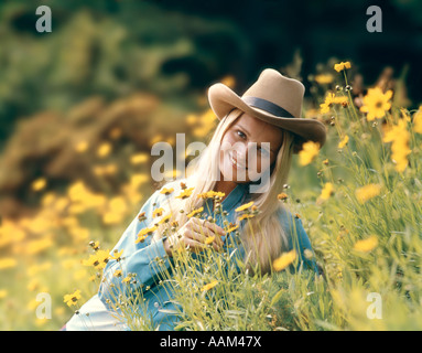 1970ER-JAHRE RETRO-BLONDEN FRAU IN COWBOY-HUT SITZT IN EINEM FELD VON BUTTERCUP WILDE BLUMEN LÄCHELN Stockfoto