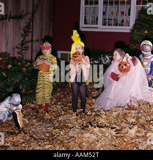 1970ER JAHRE GRUPPE VON FÜNF JUNGEN UND MÄDCHEN IN HALLOWEEN-KOSTÜMEN HÄLT TRICK OR TREAT KÜRBISSE UND TASCHEN IM VORGARTEN DES HAUSES Stockfoto