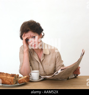 1970ER JAHREN FRAU HOLDING ZEITUNG MIT KAFFEE UND KUCHEN BESORGT SCHAUEN AUF GESICHT, BLICK IN DIE KAMERA Stockfoto