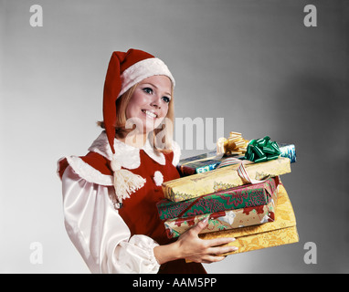 1960ER JAHRE JUNGE FRAU IN ROT UND WEIß SANTA HELFER KOSTÜM UND HUT HÄLT HAUFEN VON UMSCHLOSSENEN WEIHNACHTEN PRÄSENTIERT STUDIO Stockfoto